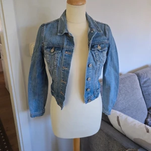 Blå croppad jeansjacka - Säljer en klassisk blå croppad jeansjacka med knappar framtill och två bröstfickor. Jackan har lång ärm och en snygg, figurnära passform. Perfekt att slänga över en t-shirt eller klänning för en cool look.