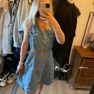 Jeansklänning från Chimi - Säljer en snygg jeansklänning i ljusblå denim. Klänningen har kort ärm, v-ringning, knappar framtill och bröstfickor. Midjan markeras med ett brunt bälte. Perfekt för dig som gillar en avslappnad men trendig stil. Säljer åt mamma från 80/90 talet. 