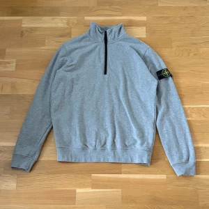 Stone Island half-zip tröja - Säljer en grå långärmad half-zip tröja från Stone Island med klassisk logotyp-patch på ärmen. Tröjan har hög krage och ribbade muddar vid ärmslut och nederkant. Perfekt för en stilren och avslappnad look. Påriktigt typ helt oanvänd 
