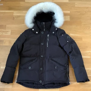  Moose Knuckles jacka - Säljer en svart dunjacka från Moose Knuckles med vit pälskant på huvan. Jackan har dragkedja, knappar och flera fickor med dragkedjor. Broderad detalj på insidan och logga på ärmen. Perfekt för kalla dagar. Kolla ”defekt” på sista bilden 