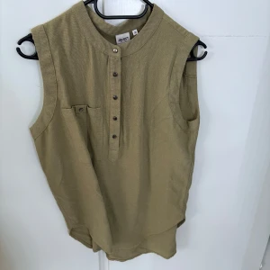 Beige ärmlös linnetopp från Object - Säljer en beige ärmlös linnetopp från Object med ståkrage och knappar framtill. Toppen har en liten bröstficka och är tillverkad i ett svalt linnematerial, perfekt för varma dagar.