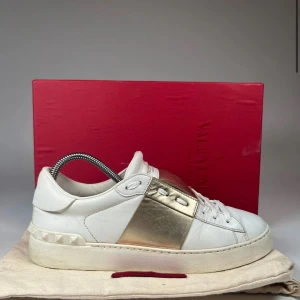 Valentino Open sneakers i vitt och guld - Snygga Valentino Open sneakers i vitt skinn med en bred guldfärgad panel på sidan. Skorna har vita snören, rund tå och platt sula med ikoniska nitar på hälen. Perfekta för dig som vill ha en lyxig och stilren look.