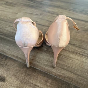 Ljusrosa klackskor med öppen tå från Cruz Pradera - Säljer ett par eleganta ljusrosa klackskor från Cruz Pradera. Skorna har öppen tå, smal klack och fina utskurna detaljer framtill. Rem runt vristen med spänne. Perfekta för dig som vill ha något extra till festen. Väll använda