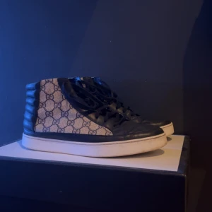 Gucci sneakers med monogram och svarta detaljer - Säljer ett par höga sneakers från Gucci med klassiskt GG-monogram i beige och mörkblått tyg. Skorna har svarta skinnpaneler och snörning framtill samt vit platt sula. Perfekta för dig som gillar lyxiga streetwear-vibbar.