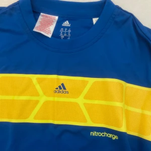 Blå t-shirt från Adidas med gul detalj - Säljer en blå Adidas t-shirt med en bred gul panel över bröstet och texten 'nitrocharge'. Klassisk rund hals och korta ärmar. Perfekt för dig som gillar sportig stil. Står xl men är mindre än så, mer som M/s