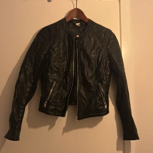 Svart skinnjacka H&M med dragkedjor - Säljer en svart skinnjacka med coola detaljer som quiltade axlar och dragkedjor vid fickorna. Jackan har lång ärm och ribbade muddar vid ärmslut. Perfekt för dig som vill ha en edgy look. Denna jacka är från H&M och är inte bara i skinn utan i annat material också som du kan se på sista bilden. 