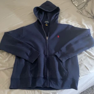 Mörkblå hoodie från Polo Ralph Lauren - Säljer en mörkblå hoodie från Polo Ralph Lauren med dragkedja och huva. Klassisk röd broderad logga på bröstet och två fickor framtill. Perfekt för en chill och stilren look.