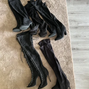 Svarta lårhöga boots med klack - Fem par svarta lårhöga boots med klack. Ett par har snörning framtill och spetsig tå, de andra har slät design och rund tå. Alla är i mocka-liknande material och har höga klackar. Perfekta för att lyfta din outfit.   