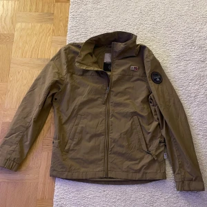 Beige vindjacka från Napapijri - Säljer en beige vindjacka från Napapijri med dragkedja framtill och två sidofickor. Jackan har hög krage, patch med logga på ärmen och norsk flagga på bröstet. Perfekt för blåsiga dagar och enkel att matcha med det mesta. Con 9/10