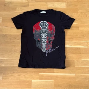 Philipp Plein Junior T-shirt  - Svart t-shirt från Philipp Plein Junior med ett stort döskalletryck i rött, grått och silver på framsidan. T-shirten har rund hals och korta ärmar. Perfekt för dig som gillar statement-plagg. Tröjan sitter som S/M. Defekter ses på sista bilden.