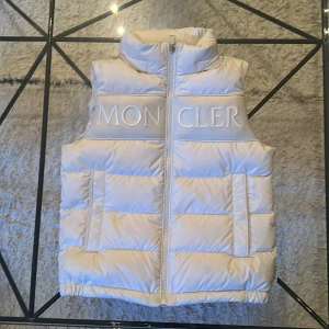 Vit dunväst från Moncler - Snygg och puffig vit dunväst från Moncler med hög krage och dragkedja framtill. Västen har två fickor på sidorna och tydlig Moncler-logga broderad över bröstet. Perfekt för lager-på-lager och en clean look. Köpte den på NK för 3800kr helt ny.