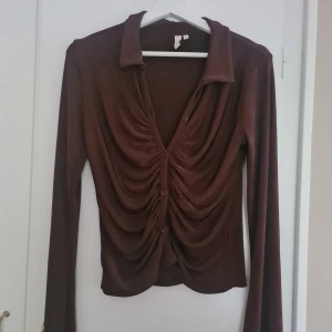 Brun långärmad blus från Nelly  - Säljer en brun långärmad blus från Nelly med snygga rynkade detaljer framtill och knappar längs hela framsidan. Blusen har krage och är tillverkad i ett mjukt, stretchigt material som sitter snyggt på kroppen.