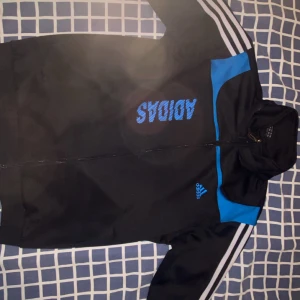 Svart träningsjacka från Adidas med blå detaljer - Säljer en svart träningsjacka från Adidas med blå detaljer och klassiska vita ränder på ärmarna. Jackan har dragkedja framtill och Adidas-logga på bröstet. Perfekt för sport eller chill.