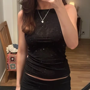 Svart glittrig linnetopp - Snygg svart linnetopp med glittriga detaljer och hög halsringning. Perfekt för att lyfta en outfit med lite extra bling. Toppens material är stretchigt och sitter tight mot kroppen.