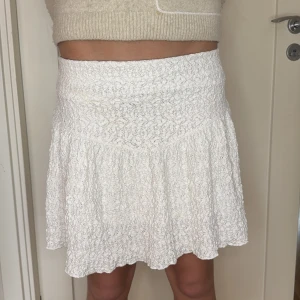 Vit spetskjol med volang - Säljer en söt vit kjol med bred midja och volangkant. Kjolen har ett fint spetsmönster och är lätt utsvängd för en feminin look. Perfekt att matcha med en enkel topp.