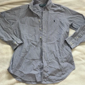 Randig skjorta från Ralph Lauren - Klassisk blå- och vitrandig skjorta från Ralph Lauren med broderad logga på bröstet. Skjortan har lång ärm, knappar framtill och klassisk krage. Säljer skjortan eftersom jag tycker den är lite för stor för mig. Det finns inga defekter på den och den är i mycket bra skick. 