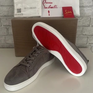 Gråa sneakers från Christian Louboutin - Säljer ett par gråa sneakers från Christian Louboutin med den ikoniska röda sulan. Skorna har snörning, rund tå och är tillverkade i mocka. Perfekta för dig som vill ha en stilren och lyxig look.
