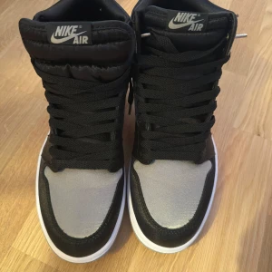 Nike Air Jordan 1 Retro High OG Silver/Black - Snygga Nike Air Jordan 1 Retro High OG i svart och silver med vita sulor. Skorna har klassisk hög siluett, svart snörning och ikoniska Air Jordan-loggan på sidan. Perfekta för dig som vill sticka ut med stilrena sneakers.