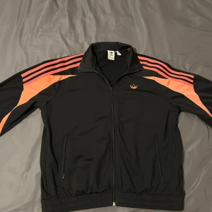 Svart Adidas track jacket med orange ränder - Säljer en svart Adidas track jacket med klassiska orange ränder längs ärmarna och dragkedja framtill. Jackan har ståkrage, två fickor med dragkedja och ribbade muddar. Perfekt för en sportig och avslappnad stil.