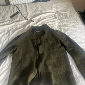 Olivgrön cardigan jacka från j.lindeberg - Säljer en olivgrön cardigan jacka från j.lindeberg med dragkedja framtill och en bröstficka. Jackan har en enkel och stilren design, perfekt för lager-på-lager. Passar dig som gillar minimalistisk stil.