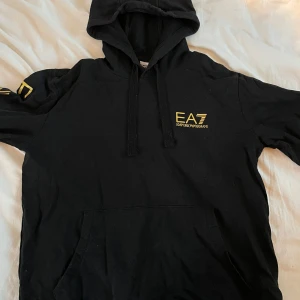Svart hoodie från Emporio Armani EA7 - Säljer en svart hoodie från Emporio Armani EA7 med guldiga loggor på bröstet och längs ena ärmen. Tröjan har huva med dragsko och en stor magficka framtill. Perfekt för dig som gillar stilrena och exklusiva plagg.
