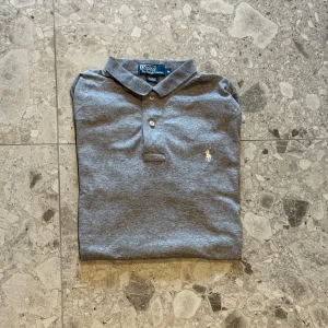 Grå pikétröja från Polo Ralph Lauren - Klassisk grå pikétröja från Polo Ralph Lauren med korta ärmar och krage. Matrialet är bomull och nypriset ligger runt cirka 1500kr