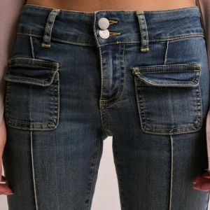 Blå jeansbyxor med fickor fram - Säljer ett par blå jeansbyxor från Nelly med två stora fickor framtill och markerade sömmar längs benen. Byxorna är lågmidjade och stängs med dubbla knappar och dragkedja. Stl 38