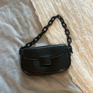 Svart axelväska med kedjehandtag - Snygg svart axelväska i glansigt syntetmaterial med bred kedja som handtag. Väskan har en stilren design med lock och fyrkantigt spänne framtill. Perfekt accessoar för att lyfta din outfit.