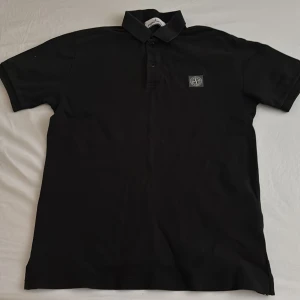 Svart pikétröja från Stone Island - Säljer en svart pikétröja från Stone Island med klassisk krage och knappslå. Tröjan har korta ärmar och en broderad logotyp på bröstet. Perfekt för en stilren och avslappnad look.