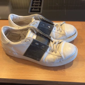 Vallentino open - Säljer ett par vita sneakers från Vallentino med bred svart rem över sidan. Skorna har snörning och rund tå. Klassisk och stilren design med platt sula och diskret logga på plösen. Skorna är I slitet men ändå okej skick så säljs därmed billigt. Hör av er vid funderingar