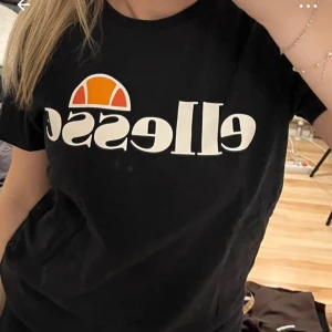 Svart t-shirt från Ellesse - Svart t-shirt från Ellesse med stort vitt tryck och logga framtill. Klassisk rund halsringning och korta ärmar. Perfekt för en sportig och avslappnad stil.