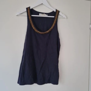 Svart linnetopp med gulddetaljer - Snygg svart linnetopp med breda axelband och dekorativa guldfärgade pärlor runt ringningen. Perfekt att styla till både jeans och kjol för en trendig look.