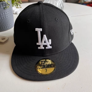 Svart LA-keps från New Era 59FIFTY - Säljer en svart keps från New Era 59FIFTY med LA-logga broderad i vitt framtill. Klassisk modell med rak skärm och broderade ventilationshål. Tillverkad i polyester och passar perfekt för dig som gillar sportig stil. Passar folk som har huvudstorlek 55,8cm! Hör av er vid minsta fundering mvh Theo