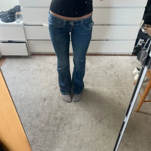 Blå bootcut jeans med låg midja - Säljer ett par klassiska blå bootcut jeans med låg midja. Jeansen har fickor, dragkedja och knapp framtill samt snygga sömdetaljer på bakfickorna. Perfekta för en avslappnad och trendig look.