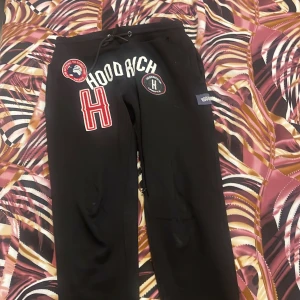 Svarta mjukisbyxor från Hoodrich med broderade detaljer - Svarta mjukisbyxor från Hoodrich med stora broderade loggor och text på framsidan. Byxorna har dragsko i midjan och flera patchar i rött, vitt och blått. Perfekta för en avslappnad streetwear-look.