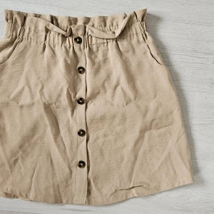 Beige kjol med knappar (dekoration)och resår - Snygg beige kjol med knappar framtill Kjolen har fickor på sidorna och är i en mjuk bomullsblandning. Perfekt för en avslappnad och trendig look. Kjolen är bara använd några enstaka gånger. Har haft en svart och använt den tupp dagligen, köpte därför en beige också, men svårt med att hitta snygga plagg till. Den 