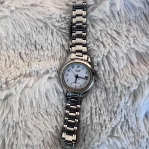 Silverfärgad vintageklocka från Citizen - Stilren klocka från Citizen med silverfärgad länkarmband och vit urtavla. Klockan har tydliga svarta siffror, datumvisning och klassisk rund form. Måste byta batteri