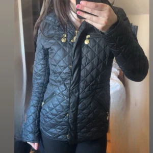 Svart quiltad jacka med guldiga detaljer - Snygg svart quiltad jacka med guldiga knappar och dragkedja. Jackan har lång ärm, hög krage och figurnära passform. Perfekt för kyligare dagar och enkel att matcha med olika outfits.