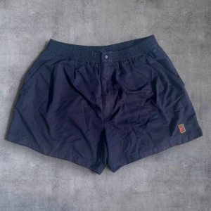 Mörkblå shorts från Nike - Mörkblå shorts från Nike med elastisk midja och knapp framtill. Shortsen har sidofickor, bakficka med knapp och ett broderat Nike-märke på vänster ben. Perfekta för sommardagar vid vattnet.