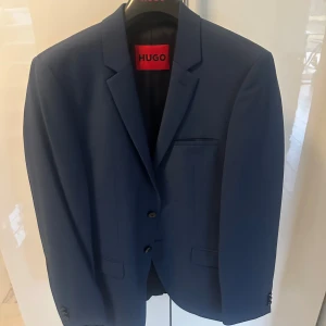 Blå kostym från Hugo Boss  - Säljer en stilren blå kostym från Hugo Boss bestående av kavaj och byxor, levereras i original röd kostympåse. Tillhörande svart bälte med röd insida ingår. Perfekt för bal eller festliga tillfällen.