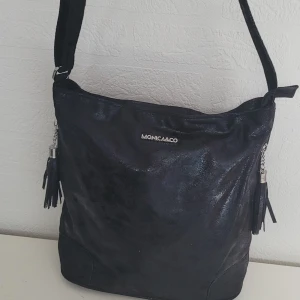  Helt ny Svart axelväska från MONICA&CO -  Helt ny Snygg svart axelväska från MONICA&CO med glansig finish och tofsdetaljer på sidorna. Väskan har dragkedja upptill och ett extra fack med dragkedja på framsidan. Perfekt för att bära dina viktigaste saker med stil.