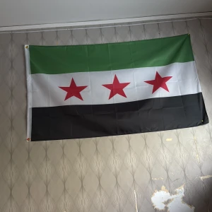 Syriska oppositionsflaggan - Säljer en flagga med tre horisontella ränder i grönt, vitt och svart samt tre röda stjärnor i mitten. Flaggan är rektangulär och tillverkad i tyg, perfekt att hänga på väggen eller använda vid evenemang.