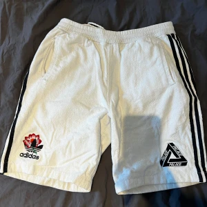 Vita Adidas x Palace shorts - Säljer ett par vita shorts från Adidas och Palace med svarta ränder på sidorna, broderade loggor fram och bak samt dragsko i midjan. Shortsen har två sidofickor och är tillverkade i frotté-material. Perfekta för sommaren! Litet hål vid ena fickan och lite sönder där man knyter men funkar fortf fint. Sjukt sällsynta.