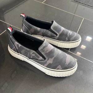 Camo slip-on sneakers - Säljer ett par slip-on sneakers från New Look med grått camouflage-mönster och vit, platt sula. Skorna har elastiska paneler på sidorna och en liten röd, vit och svart detalj på hälen. Perfekta för en avslappnad stil.