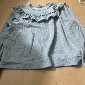 Grå tröja med volang från H&M - Säljer en ljusgrå långärmad tröja från H&M med volangdetalj över bröstet. Tröjan har rund halsringning och är tillverkad i mjukt bomullsmaterial. Perfekt för en avslappnad och stilren look.