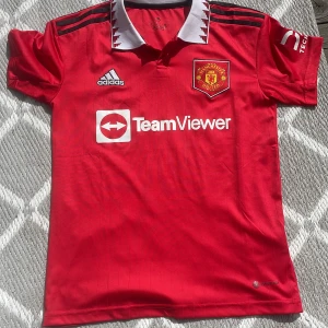 Manchester United hemmatröja 2022/23 - Röd Manchester United fotbollströja från Adidas med vit krage och svarta detaljer. Broderad klubbmärke på bröstet, Adidas-logga och sponsortryck. Klassisk pikékrage med mönster. Perfekt för fans!