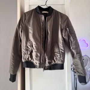 Brun bomberjacka med svarta detaljer - Säljer en stilren brun bomberjacka med svarta muddar och krage. Jackan har dragkedja framtill, fickor med tryckknappar och en ärmficka med dragkedja. Perfekt för dig som vill ha en klassisk och enkel look.