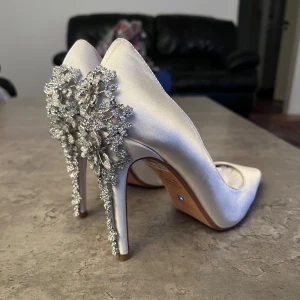 Vita pumps med glittriga dekorationer - Eleganta vita pumps i satin med spetsig tå och hög klack. Baksidan av klacken är dekorerad med stora, glittriga blomliknande smycken i silverfärg. Perfekta för speciella tillfällen.