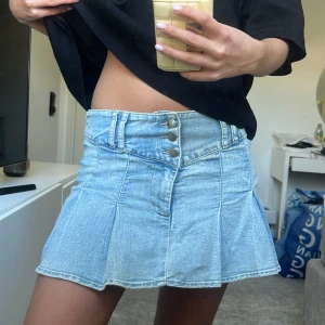 Ljusblå jeanskjol med knappar - Säljer en ljusblå jeanskjol med hög midja och tre synliga knappar framtill. Kjolen har en kort och utsvängd modell som ger en snygg siluett. Perfekt för dig som gillar en trendig och avslappnad stil.