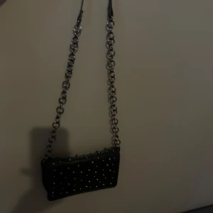 Svart axelväska med nitar och kedja - Cool svart axelväska med silverfärgade nitar och en grov kedja som axelrem. Väskan har en rektangulär form och passar perfekt för att lyfta en outfit med lite edge. Perfekt storlek för det viktigaste!